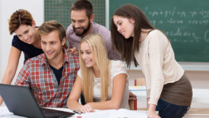 Successful team of diverse young male and female students in the classroom smiling with delight as they read the outcome of their project on a laptop computer